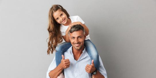 Glücklicher Vater mit seiner Tochter auf den Schultern, beide lächeln in die Kamera, vor grauem Hintergrund.