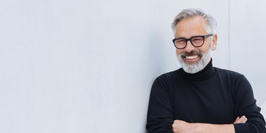 Älterer Mann mit grauem Haar, Bart und Brille steht lächelnd mit verschränkten Armen vor einer hellen Wand, trägt einen schwarzen Rollkragenpullover.
