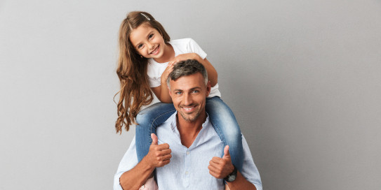 Glücklicher Vater mit seiner Tochter auf den Schultern, beide lächeln in die Kamera, vor grauem Hintergrund.