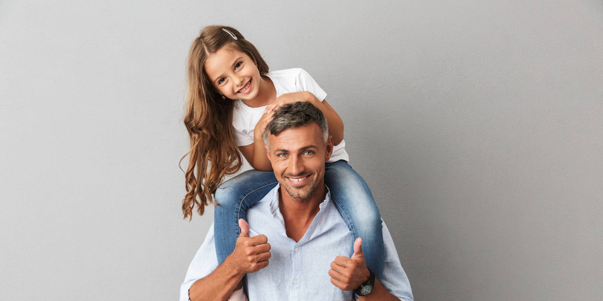Glücklicher Vater mit seiner Tochter auf den Schultern, beide lächeln in die Kamera, vor grauem Hintergrund.