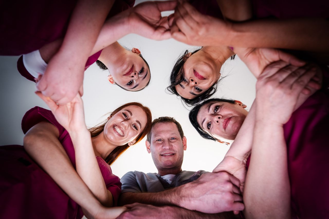 Fünf medizinische Fachkräfte bilden einen Kreis und schauen lächelnd nach unten in die Kamera, während sie sich gegenseitig an den Händen halten.