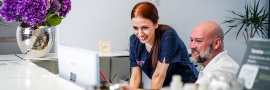 Mitarbeiterin in medizinischer Kleidung und ein Patient schauen gemeinsam auf einen Bildschirm an der Rezeption einer modernen Zahnarztpraxis.