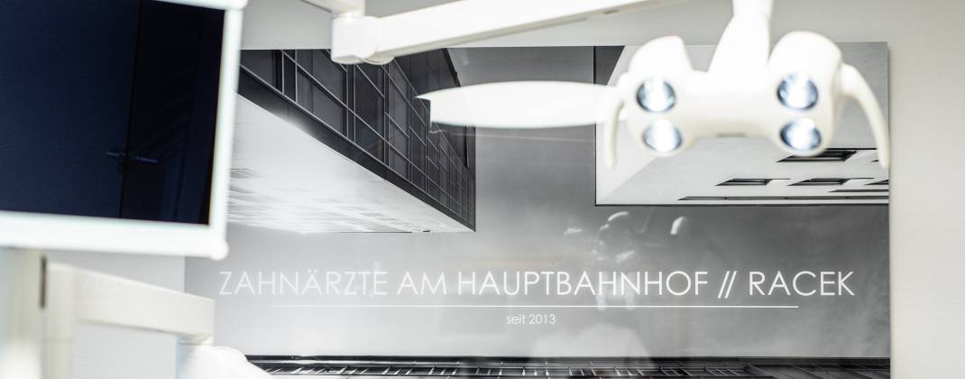 Wandbild mit moderner Architektur und dem Schriftzug „Zahnärzte am Hauptbahnhof // Racek – seit 2013“ in einer Zahnarztpraxis, im Vordergrund ein Teil der Behandlungseinheit.
