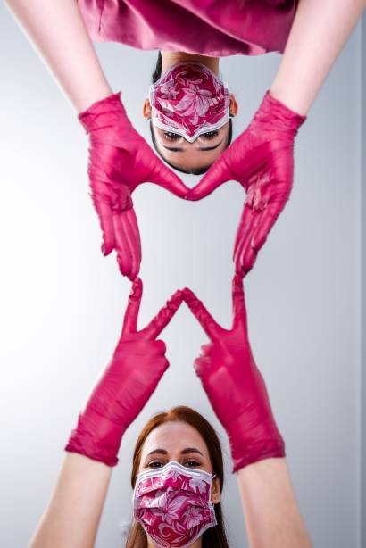 Zwei Frauen mit gemusterten Masken und pinken Handschuhen formen gemeinsam mit ihren Händen ein Herzsymbol in Richtung Kamera.