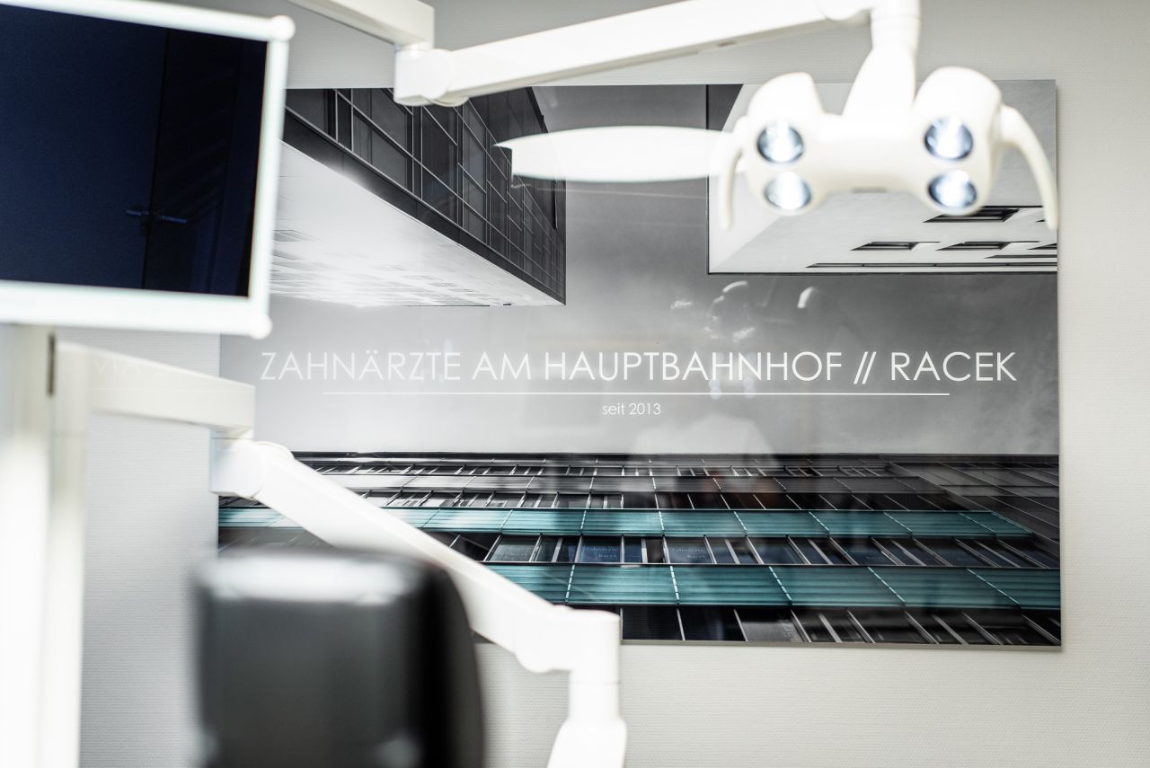 Wandbild mit moderner Architektur und dem Schriftzug „Zahnärzte am Hauptbahnhof // Racek – seit 2013“ in einer Zahnarztpraxis, im Vordergrund ein Teil der Behandlungseinheit.