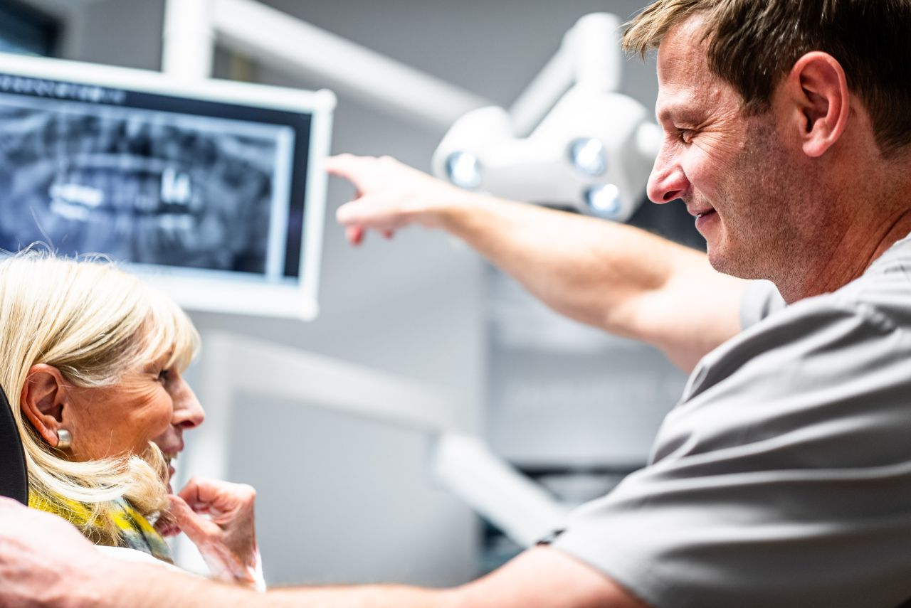 Zahnarzt zeigt einer älteren Patientin ein Röntgenbild auf einem Monitor und erklärt die Diagnose.
